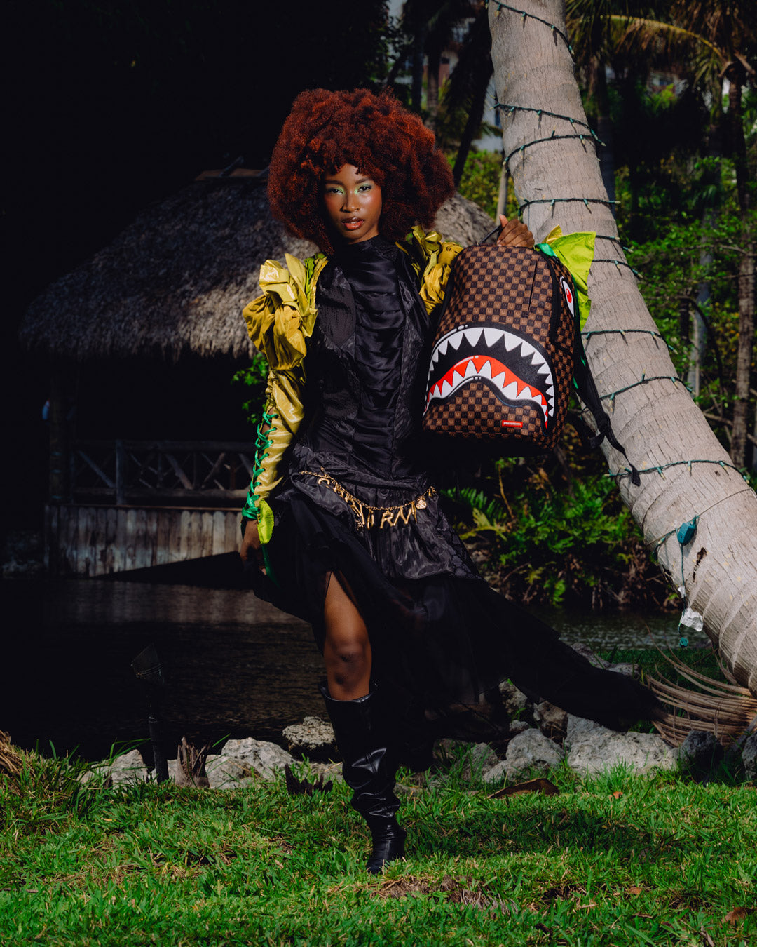 Backpack with shark-mouth graphic held by a person beside a palm-lined outdoor area