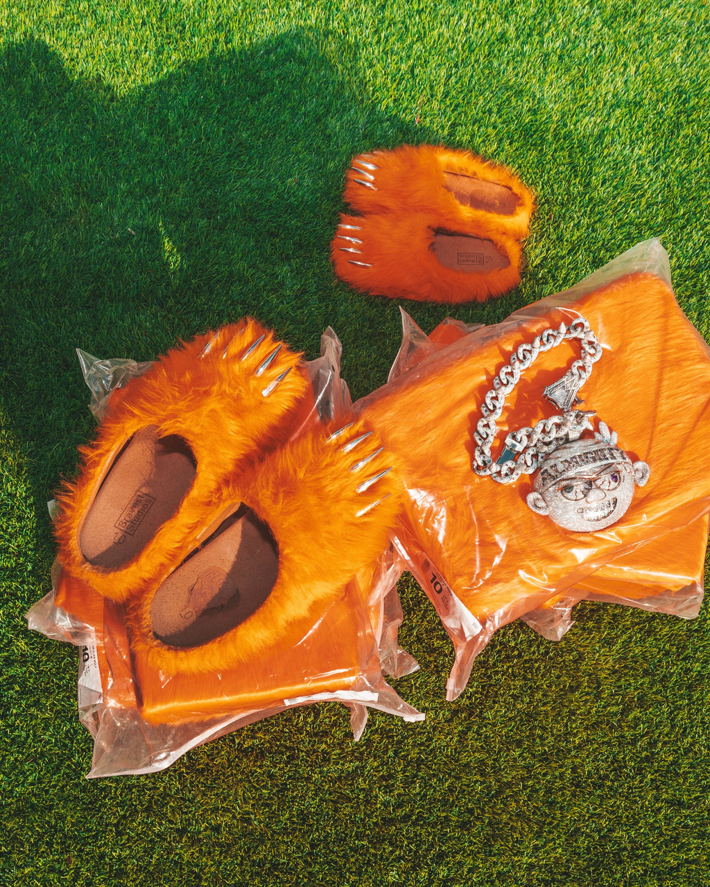 Orange plush bear-claw slippers in clear bags on grass, with silver chain charm.