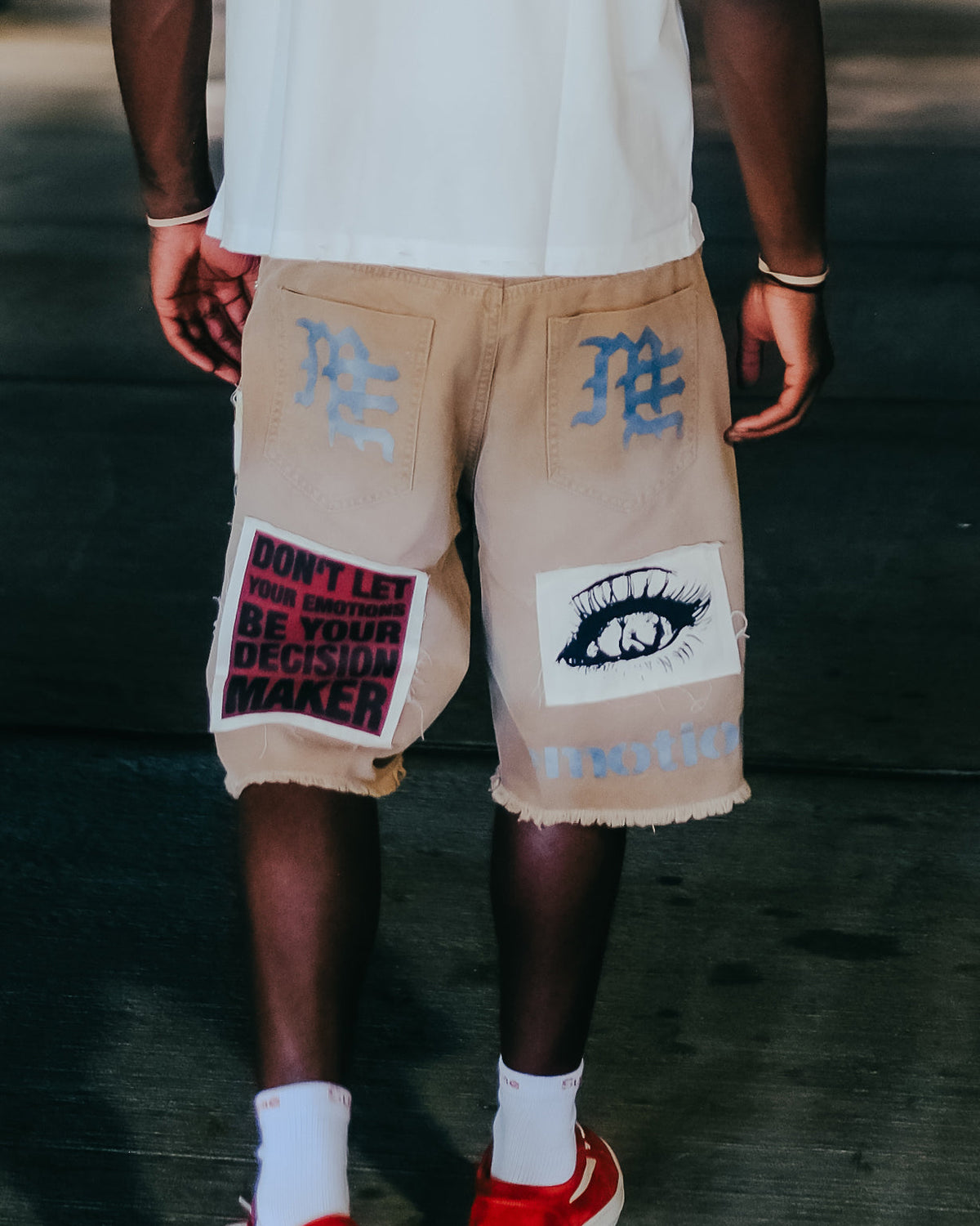 Men's brown carpenter shorts with graphic eye patch and motivational text, frayed hem