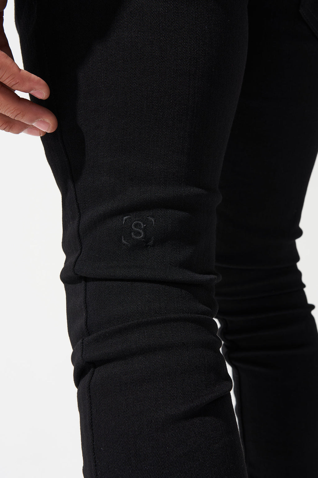 Men's dark jeans close-up on knee with hand, stacked fabric folds, and S logo