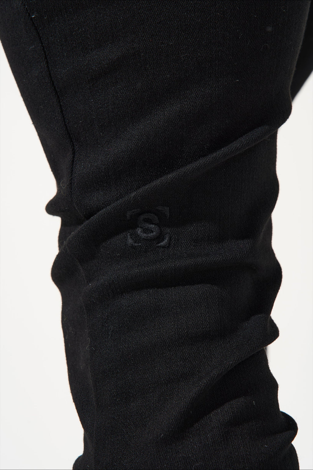 Men's dark jeans close-up of knee cuff with embossed S logo on wrinkled fabric