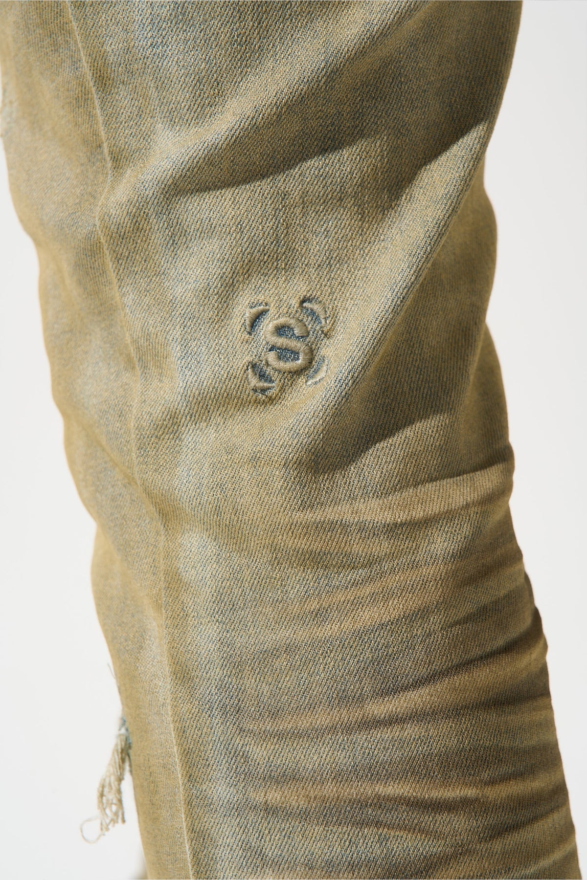Close-up of men's muted olive jeans with stitched logo mark on the thigh and frayed distress