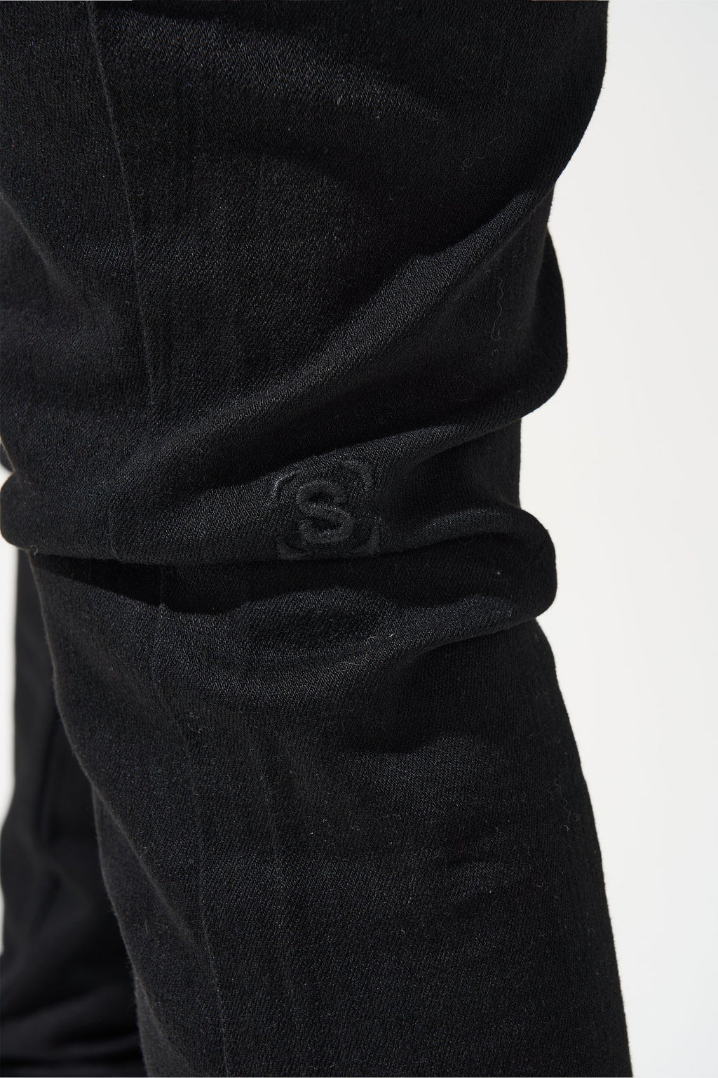 Men's dark jeans side view of knee area with S-stamp logo on cuff