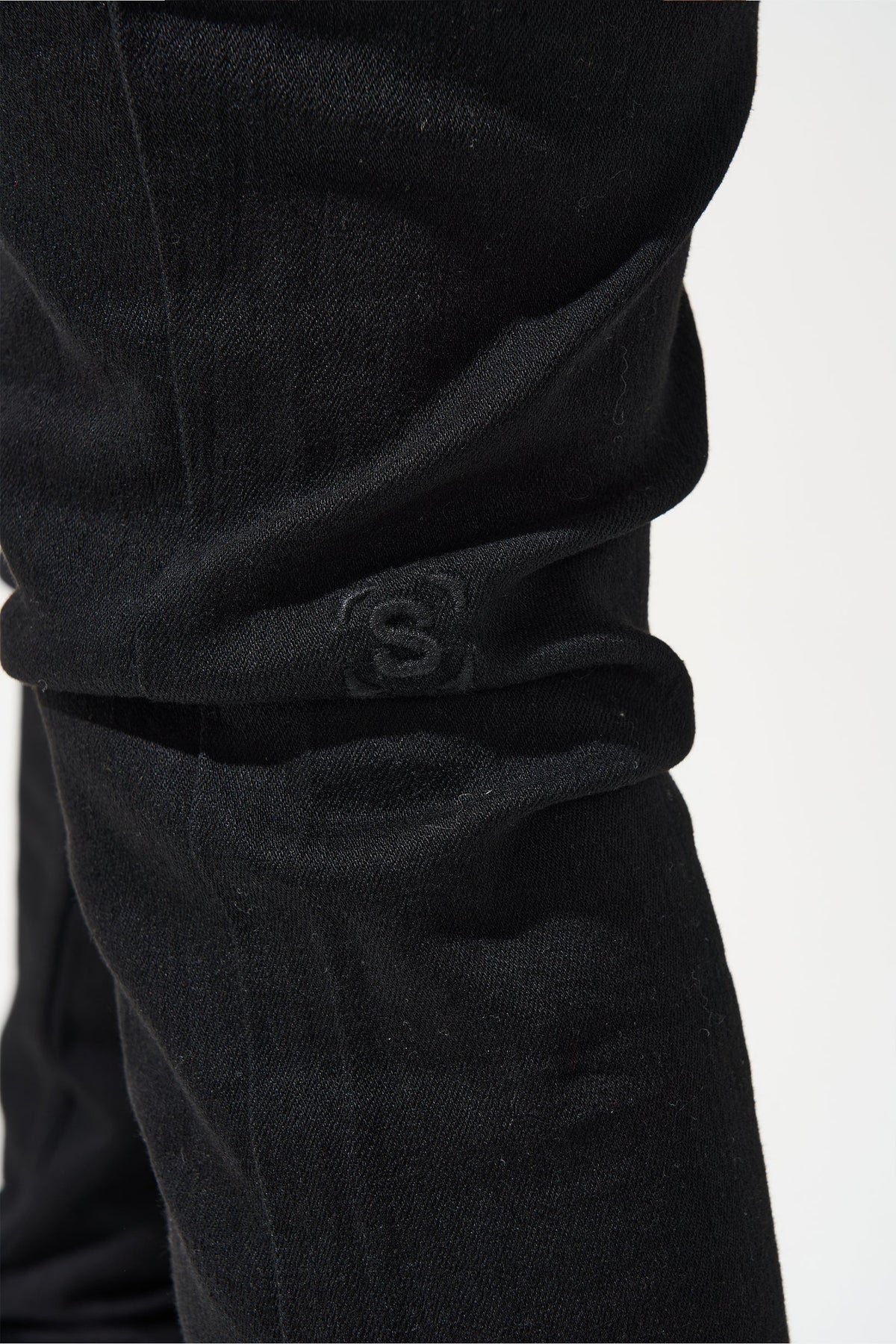 Men's dark jeans side view of knee area with S-stamp logo on cuff